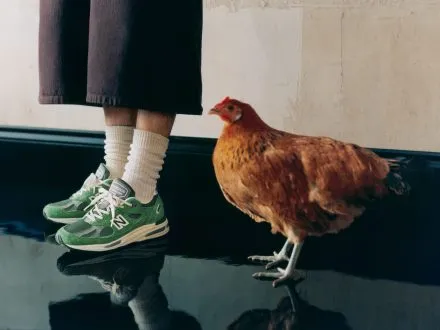 A cropped image of an unidentified person wearing trousers with green New Balance 991v2 shoes on their feet with a hen standing next to them on dark green laminated floor