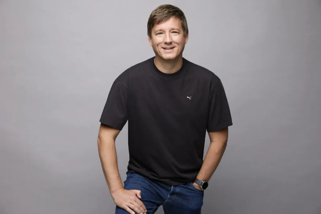 Puma COO Andres Hubert wearing black top and blue jeans smiling and posing in front of grey background
