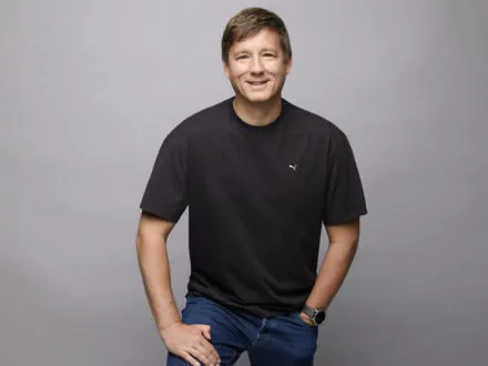 Puma COO Andres Hubert wearing black top and blue jeans smiling and posing in front of grey background