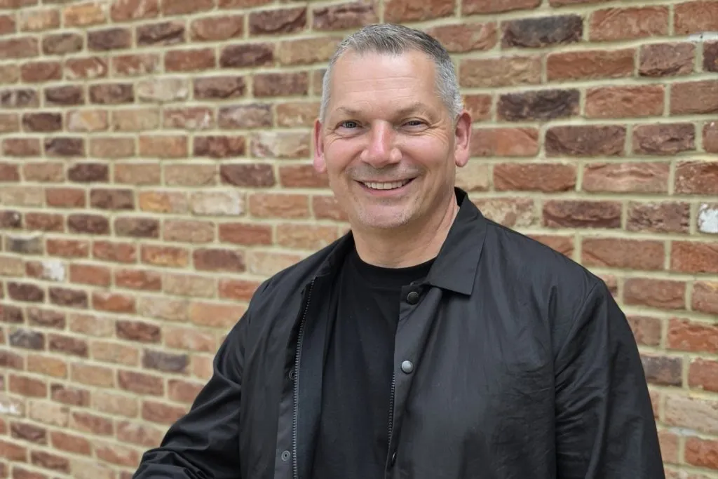 Kieron Birch standing against brick wall smiling wearing black t-shirt and jacket